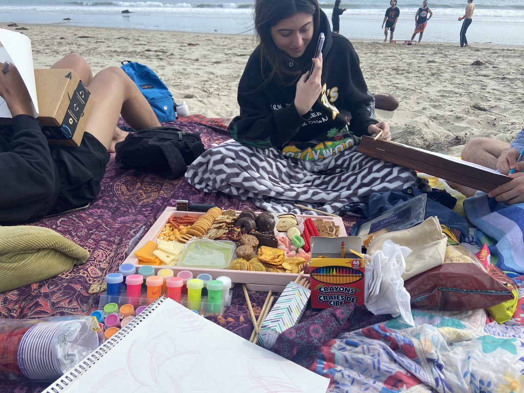 A group of three are sitting on a tapestry on the beach. There is art supplies and charcuterie board filled with cookies, crackers, cheeses, and treats on the tapestry. Bags of chips and other art supplies like paper, colored penciles, cups are on the tapestry. A young woman in a black hoodie wearing a blanket is sitting on the tapestry in the center and taking a photo of the young man on the right. The young man on the right's fingers and legs are in the picture and is drawing on a paper ontop of a folded chess board. Another young man is leaning on a pillow and using an amazon box to draw on a piece of paper. There are four young men playing spike ball on the beach in the background.