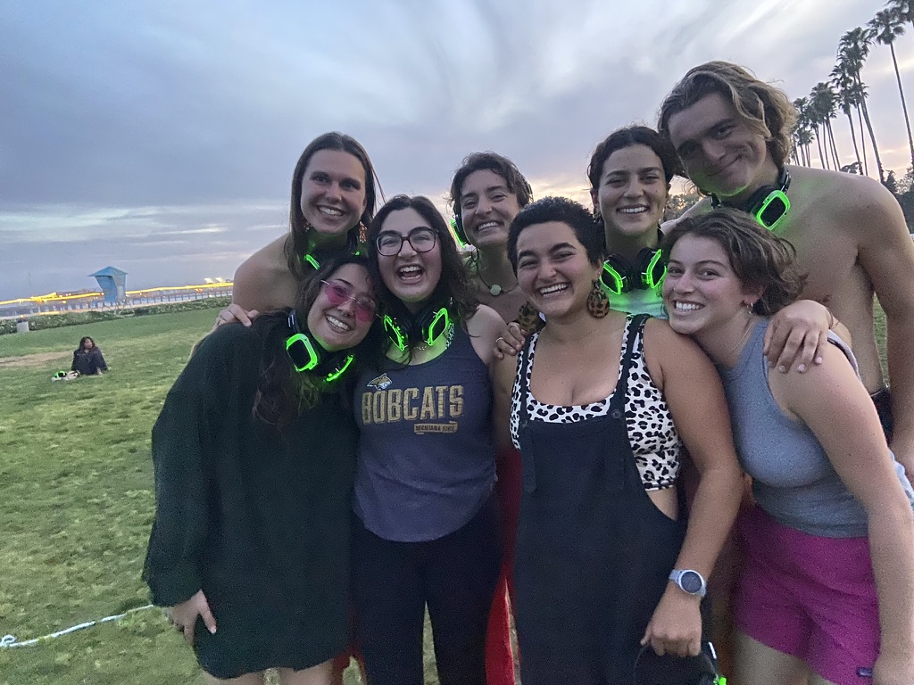 A group of 8 smiling young adults wearing neon green silent disco headphones around their necks. They are wearing a mixture of different tank tops, shorts, and tees for working out. There are palm trees, a life guard tower, the pier with string lightsm and a person sitting in the grass in the background.