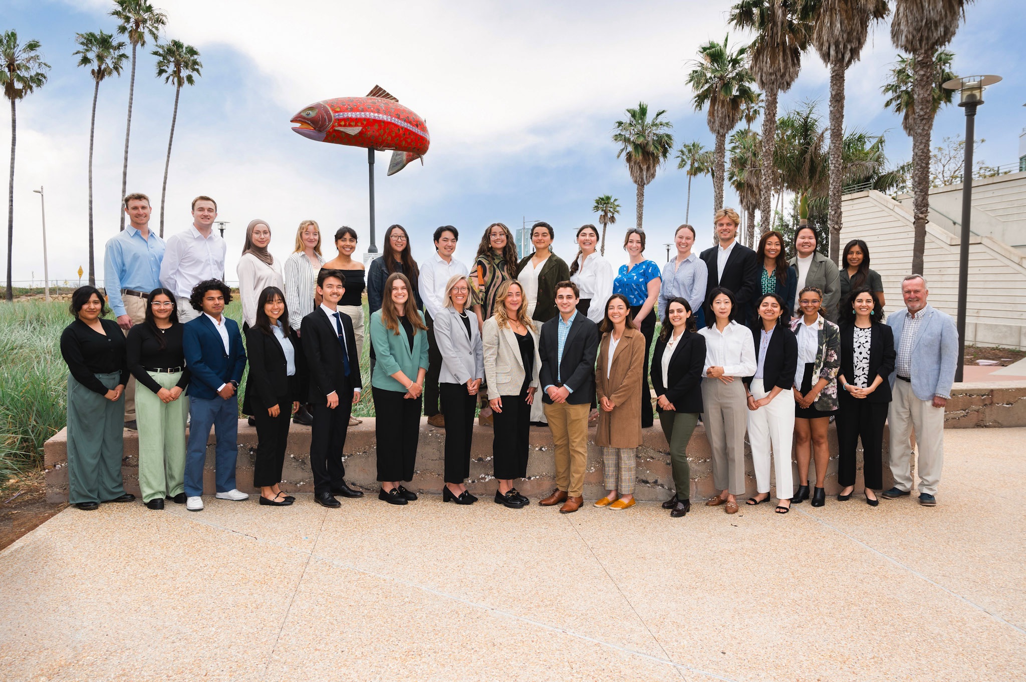 31 professionally dressed, smiling, individuals are split into two rows. There are 29 MEDS students, one professor, and one dean (both are on the bottom right side). There are palm trees and a big red fish sculpture attached to a pole in the background.